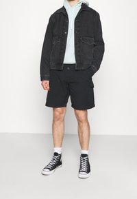 Chaqueta de mezclilla negra con dos bolsillos frontales sobre una sudadera verde clara, combinada con pantalones cortos cargo negros y zapatillas altas negras.