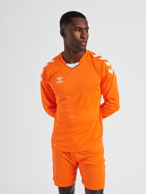Homme debout, les mains dans le dos, portant un maillot de sport orange à manches longues et un short avec des détails en chevrons blancs sur les épaules.
