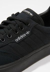 Svarta canvas sneakers med platta svarta snörningar, metallöglor, texturerad gummisula och silverfärgad Adidas-logotyp på sidan och klappen.