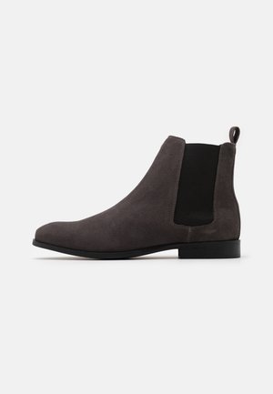 Mørkegrå ruskind Chelsea-støvle med sorte elastiske sidepaneler, trækflap bagpå, lav hæl og spids tå på hvid baggrund.