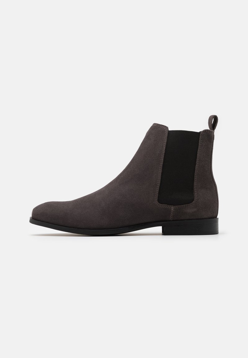 Botte Chelsea en daim gris foncé avec panneaux latéraux élastiques noirs, languette à l’arrière, talon bas et bout pointu sur fond blanc.
