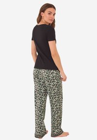 Zwarte top met korte mouwen gecombineerd met groene bloemenprintbroek. De broek heeft een patroonaccent aan de zoom. Het model staat blootsvoets, met de zijkant naar de camera gekeerd.