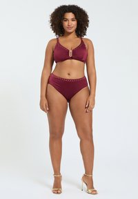 Mujer de pie con bikini burdeos con tachuelas doradas y sandalias doradas de tacón alto sobre fondo blanco.