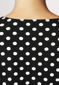 Svart tygtop med ett prickigt mönster av jämnt fördelade vita cirklar. U-ringningen är bred och texturen verkar mjuk.