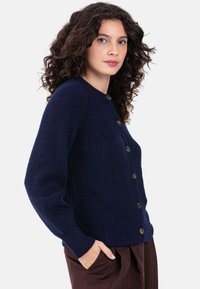 Cardigan en tricot bleu marine avec un col rond, de gros boutons marron et des poignets côtelés. La texture semble douce et chaude.