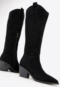 Černé semišové kozačky ke kolenům s špičatými špičkami, dekoračním prošíváním a bočním zipem. Mají robustní podpatek a poutko na horní části.
