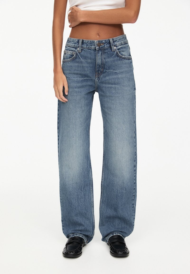 Femme portant un jean bleu taille haute, coupe ample, des mocassins noirs et un débardeur court blanc, debout devant un fond blanc.