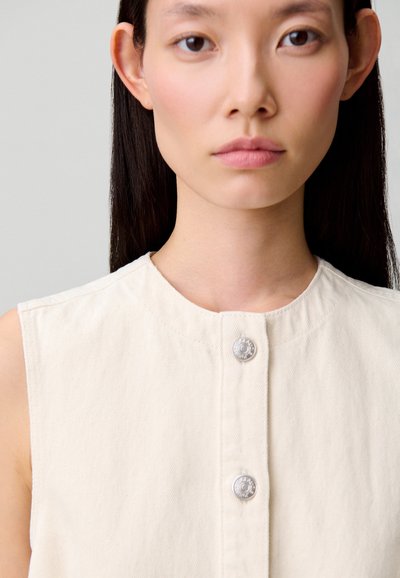 Top sin mangas de color beige claro hecho de tela texturizada, con un cuello redondo y dos botones de presión plateados en la parte delantera.