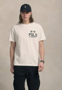 Νεαρός άνδρας με μαύρο καπέλο, λευκό μπλουζάκι Polo Motorsport, μαύρο cargo παντελόνι και ρολόι στον καρπό, στέκεται μπροστά σε ουδέτερο φόντο.