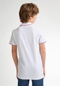 Valkoinen polo-paita, jossa on kaulus, jossa on punaiset ja siniset reunukset kauluksessa ja hihoissa. Valmistettu sileästä kankaasta, sovitettuna ja lyhyillä hihoilla.