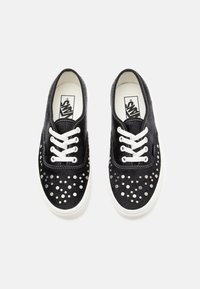 Black suede shoes with white laces feature a round studded pattern on the toe area and a white rubber sole. Classic skate shoe design.