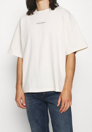 Osoba w za dużym, kremowym T-shircie z napisem "WORST BEHAVIOR" na klatce piersiowej i niebieskich dżinsach, stojąca na białym tle.