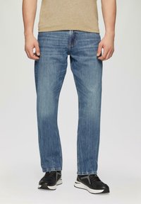 Jeans en denim bleu avec une coupe droite. Fini délavé avec une légère texture. Modèle portant des chaussures de sport noires et une chemise beige.