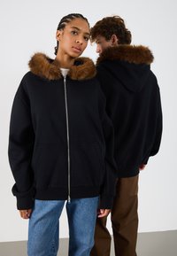 Čierny zipsový hoodie s hnedým umelým kožuchom. Obsahuje veľké predné vrecká a voľný strih. Model ho nosí s modrými džínsami.