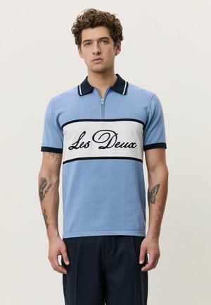 Jeune homme aux cheveux bouclés portant un polo bleu clair avec le texte « Les Deux », un col foncé et des tatouages sur les deux avant-bras.