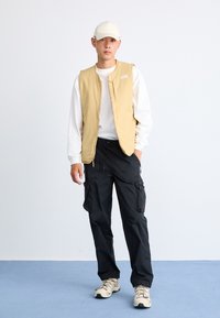 Beige sleeveless vest over a white long-sleeve shirt, paired with black cargo pants and beige shoes, set against a light background.