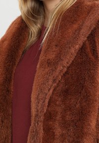 Mjuk, brun fuskpälsjacka med en bred krage och texturerad yta. Bärs över en vinröd skjorta, vilket ger ett mysigt och varmt utseende.