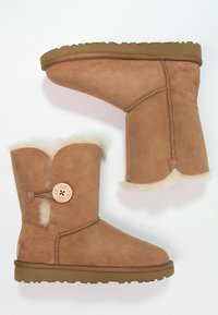 Botas de ankle de ante marrón con un cuello de piel, que presentan un botón de logo circular y una suela de goma con un patrón de relieve. Diseño de corte bajo.