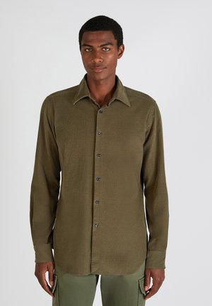 Man die rechtopstaand naar voren kijkt, gekleed in een olijfgroene overhemd met knopen en bijpassende cargo pants tegen een effen lichte achtergrond.