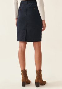 Jupe en coton bleu marine avec une fente arrière, des coutures décoratives et des accents de boutons dorés sur deux poches arrière. Assortie à des bottes à talons brunes.