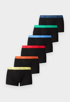 Uomo alto con slip nero con fascia blu in vita, in piedi scalzo su un tappeto, che tiene lo schienale di una sedia con struttura metallica e sedile imbottito.