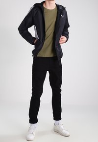 Chaqueta rompeviento negra con detalles en gris, llevada sobre una camiseta verde oliva. Pantalones negros y zapatillas blancas completan el conjunto.