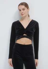Top cropped in velluto nero con maniche lunghe, caratterizzato da un design anteriore attorcigliato. Indossato con pantaloni neri in velluto abbinati a vita alta.