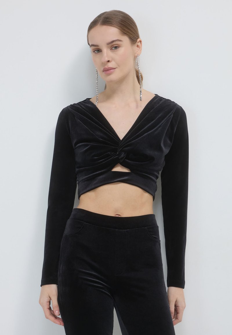 Top cropped in velluto nero con maniche lunghe, caratterizzato da un design anteriore attorcigliato. Indossato con pantaloni neri in velluto abbinati a vita alta.