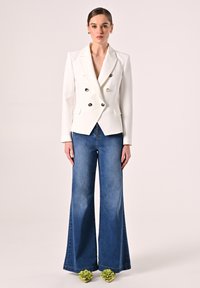 Blazer blanc à double boutonnage avec des boutons argentés, associé à un jean en denim bleu à jambes larges. Des chaussures fleuries vertes complètent le look.