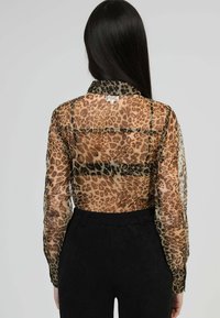 Femme aux longs cheveux bruns portant un chemisier en voile imprimé léopard et un pantalon noir taille haute, vue de dos contre un fond uni.