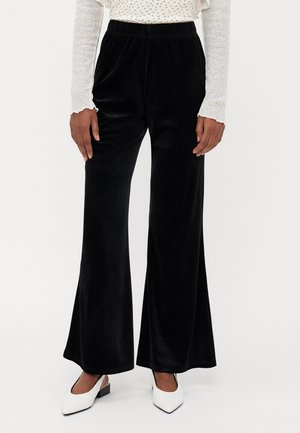Personne portant un pantalon en velours noir à jambes larges associé à des chaussures blanches à bout pointu avec un petit talon bloc, debout sur un fond uni.