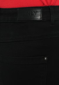 Czarne jeansy z denimu z naszywką ze skóry przyszytą do paska, posiadające tylną kieszeń z małym srebrnym guzikiem. Gładka faktura.