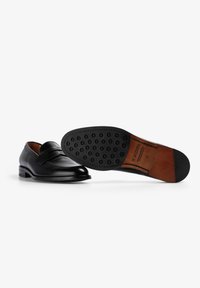 Mocasines de cuero negro con punta redonda, que presentan un detalle de correa. La suela tiene puntos de goma para proporcionar agarre y un acento marrón en contraste.