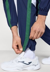 Pantalones deportivos azul marino con franjas laterales verdes, puños elásticos, detalle de cremallera en el dobladillo, combinados con zapatillas blancas y calcetines blancos.