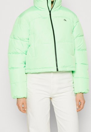 Light green puffer jacket with black zipper and small "CK" logo on chest, worn with white pants.