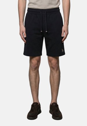 Man draagt zwarte shorts met trekkoord, beige overhemd en donkerbruine sneakers, staand tegen een witte achtergrond.