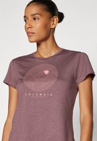 Bordeaux kortermet t-skjorte med et rosa fjellgrafikk og bølgede linjer, samt teksten "COLUMBIA SPORTSWEAR" under. Myk tekstur.