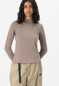 Langärmliges Rollkragenoberteil aus einem gedämpften braunen Stoff, mit schlanker Passform und kleinem dreieckigen Logo auf der Brust.