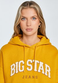 Żółta bluza z kapturem z logo z przodu, teksturowane litery "BIG STAR", kaptur na sznurku i ściągacze na mankietach. Gładki materiał w luźnym kroju.