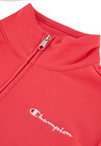Pullover rojo con cremallera y cuello alto, hecho de un tejido suave. Presenta un logo bordado en blanco de "Champion" en el pecho.