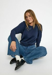 Námořnická modrá košile s dlouhým rukávem a volným střihem, v kombinaci se světle modrými kalhotami s širokými nohavicemi a černými botami. Model sedí na jednobarevném pozadí.