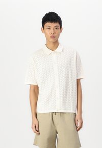 Camisa polo de manga curta em creme, com um padrão de malha em ziguezague, frente abotoada e um acabamento suave e texturizado. Usada com calções claros em caqui.
