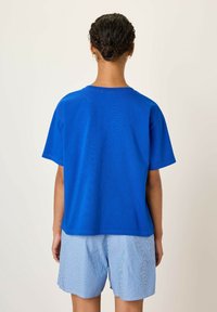 Mėlynas medvilnės marškinėliai su trumpomis rankovėmis ir apvaliu iškirpimu, derinami su šviesiai mėlynomis dryžuotomis šortais. Paprastas dizainas, kasdieniškas siluetas ir lygus tekstūra.