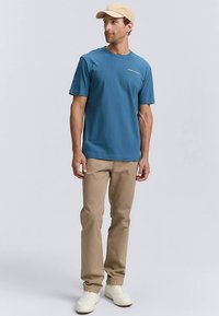 Modra bombažna majica s kratkimi rokavi in okroglim izrezom, v kombinaciji s svetlo rjavimi hlačami in belimi supergami. Majica ima diskretno blagovno znamko.