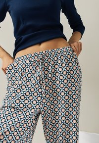 Personne portant un haut bleu marine à manches longues avec un pantalon à cordon avec des motifs comprenant des formes géométriques bleu marine, blanc et pêche, mains posées sur les hanches.