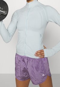 Světle modrý dlouhý zipový top s texturovanou tkaninou a otvory pro palce, spárovaný s lesklými fialovými šortkami se elastickým pasem a bočními rozparky.