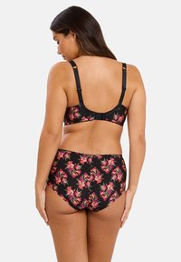 Femme vue de dos portant un soutien-gorge noir à fleurs et un slip taille haute assorti sur un fond clair uni.