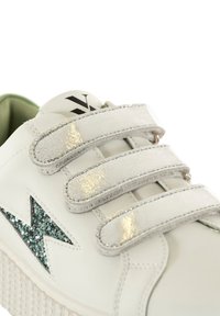 Baskets blanches avec un dessus en cuir texturé, trois bandes velcro métalliques et une étoile d'accent vert scintillant. Semelle en caoutchouc épaisse.