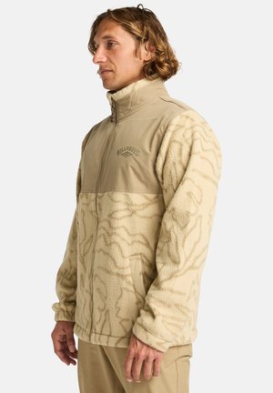 Tan fleece jakke med teksturerede cremefarvede detaljer, lynlåslukning, stående krave og lommer. Har et subtilt mønster på fleece-stoffet.