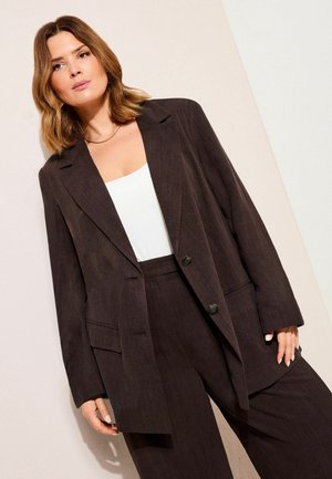 Frau mit schulterlangem hellbraunem Haar trägt einen dunkelbraunen übergroßen Blazer und passende Hose über einem weißen Oberteil, steht vor einer hellen Wand.